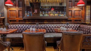 Zona de bar con asientos de cuero, paneles de madera y un cartel que dice 'Grand Hotel EST. 1928'.