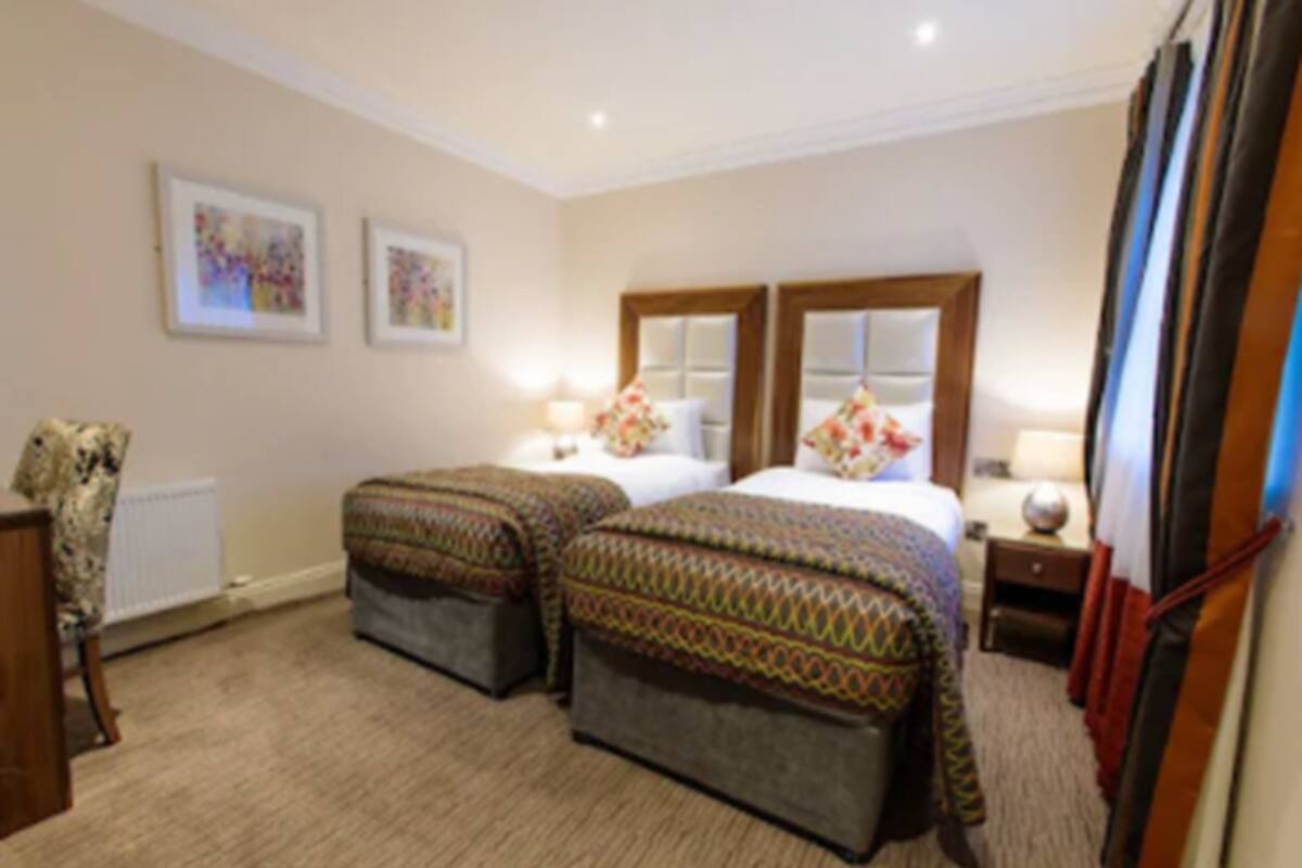 A hotel room with two beds, a dresser, a radiator, and artwork on the wall.