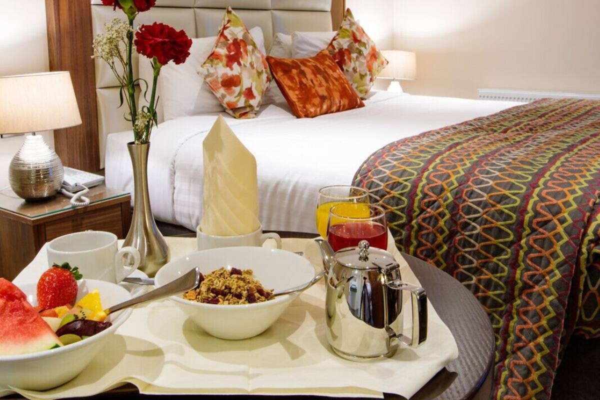 A neatly made bed with floral pillows, a bedside table with a lamp, a vase with flowers, and a breakfast tray with fruits and a teapot.