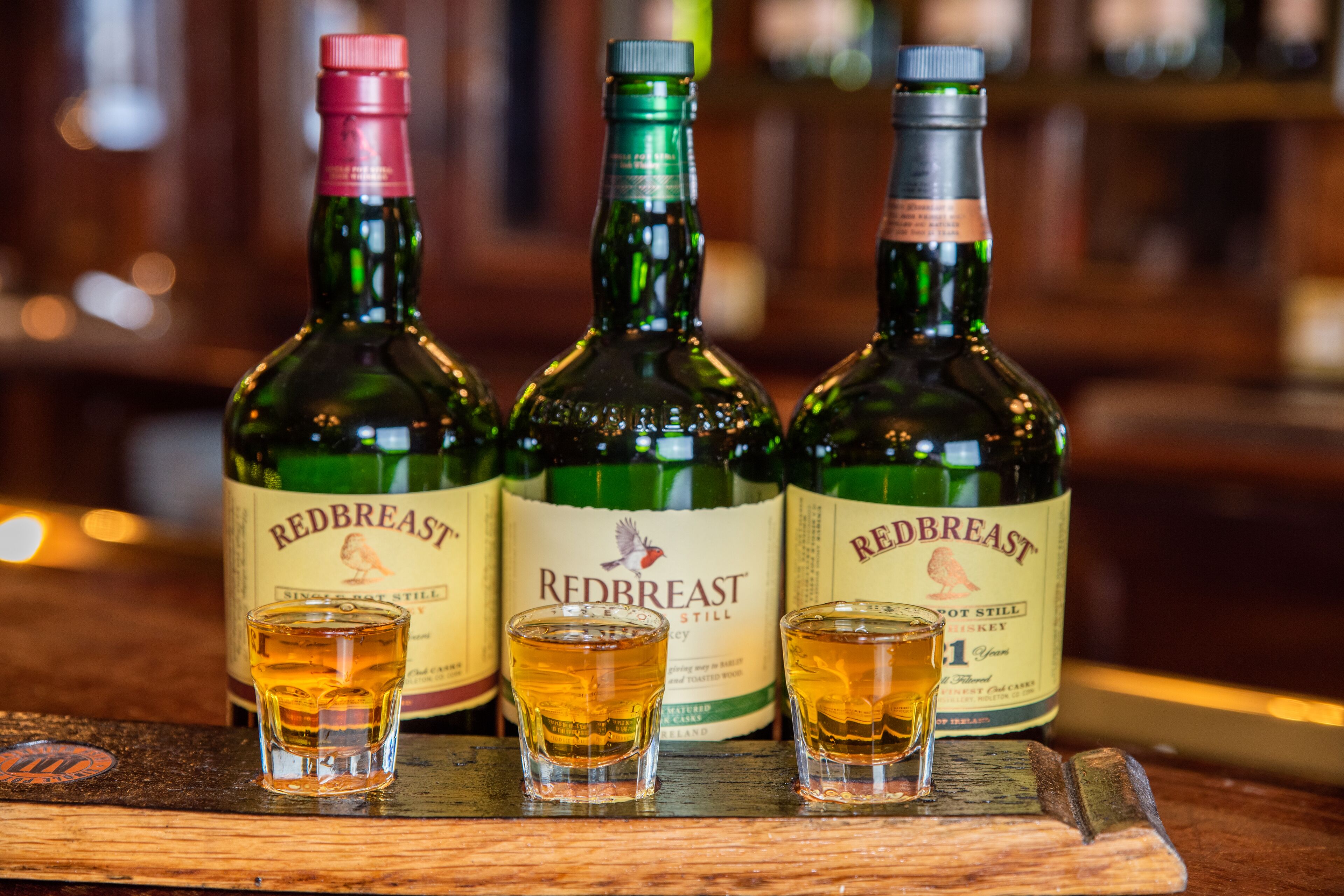Tres botellas de whisky Redbreast y dos vasos para chupito sobre una barra de madera.