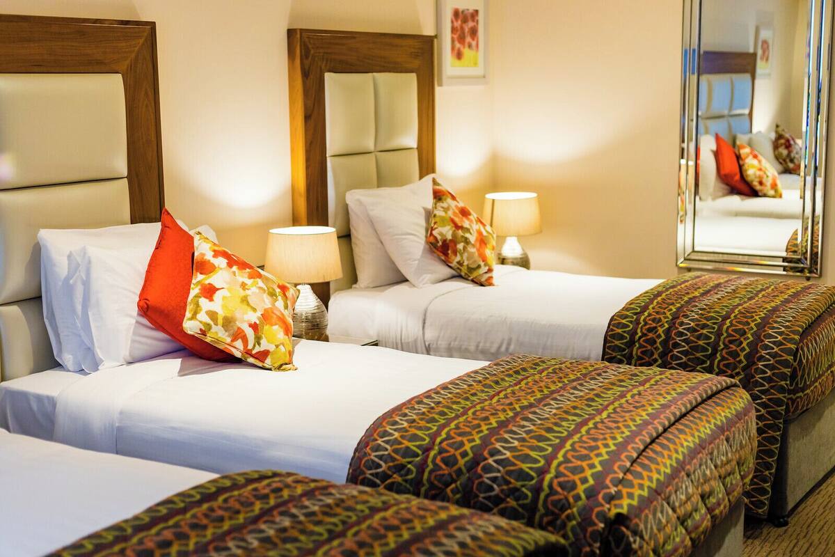 A hotel room with two beds, each with a headboard, white bedding, and decorative pillows. There are lamps on the nightstands and a mirror on the wall.