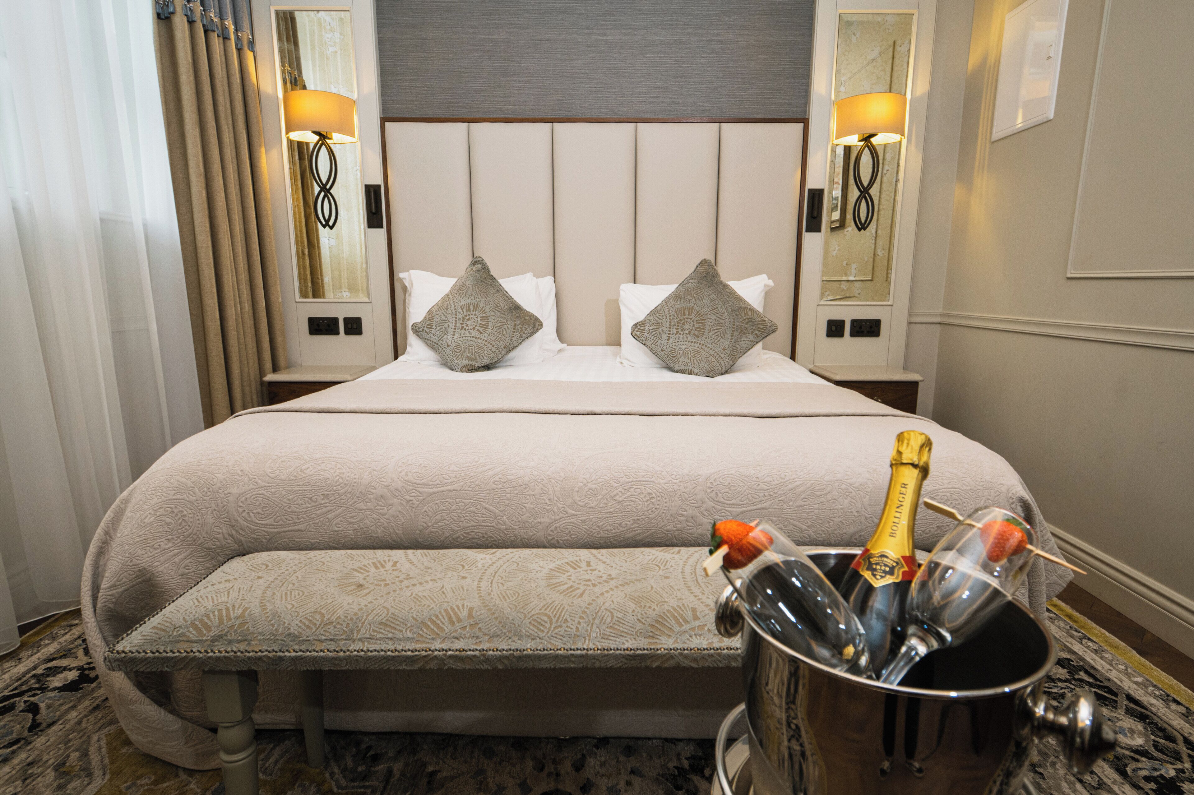A hotel room with a large bed, two bedside lamps, a nightstand, a bench, and a minibar with champagne bottles and glasses.