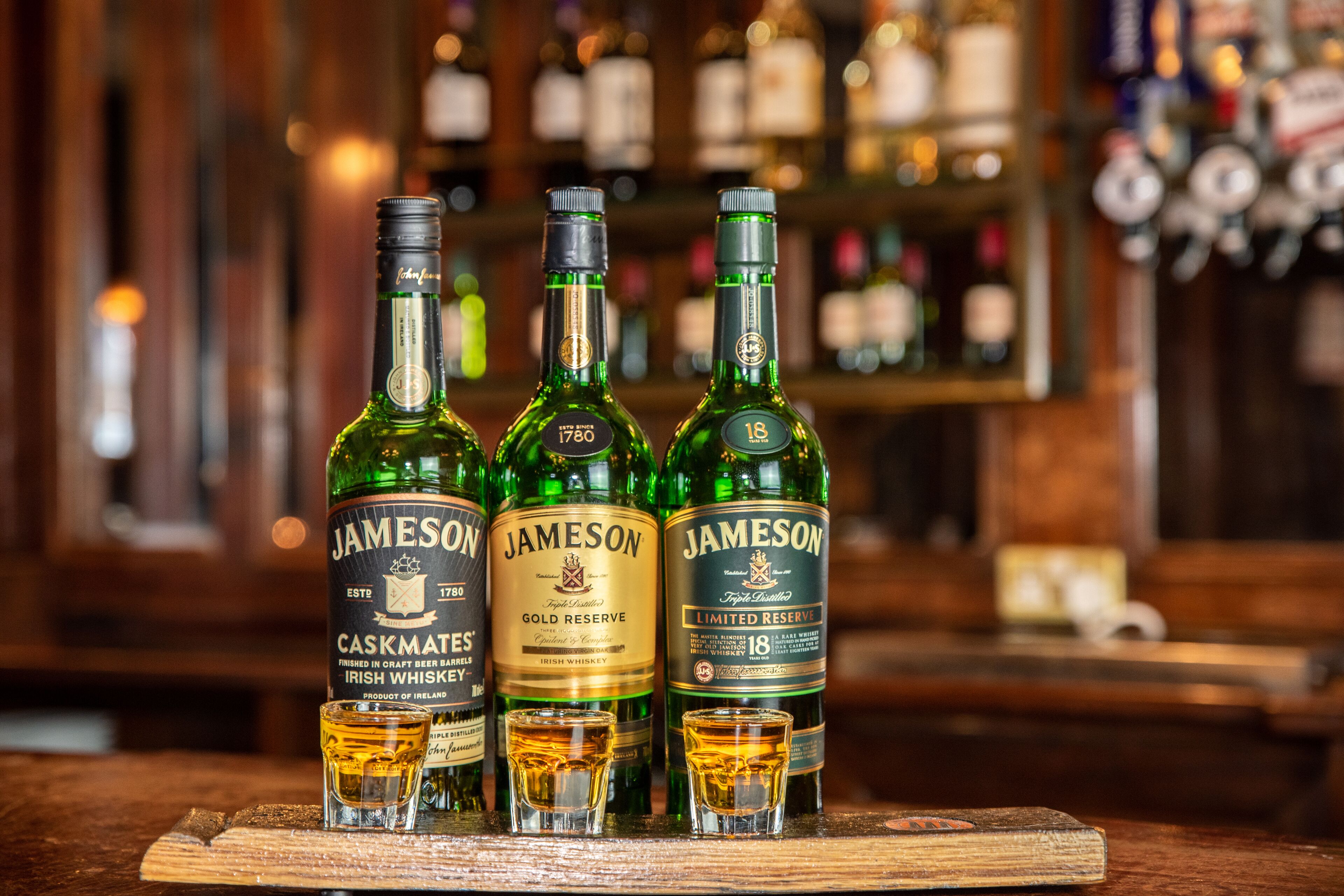 Three bottles of Jameson Irish whiskey on a wooden surface with shot glasses in front.
