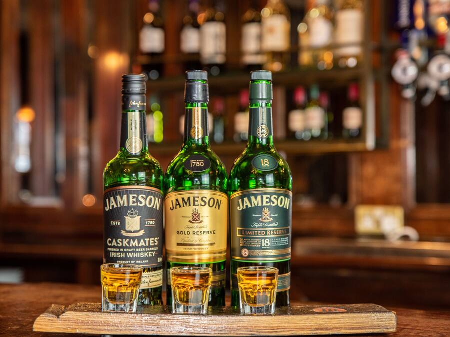 Tres botellas de Jameson Irish Whiskey sobre una superficie de madera, con copas para shots al frente.