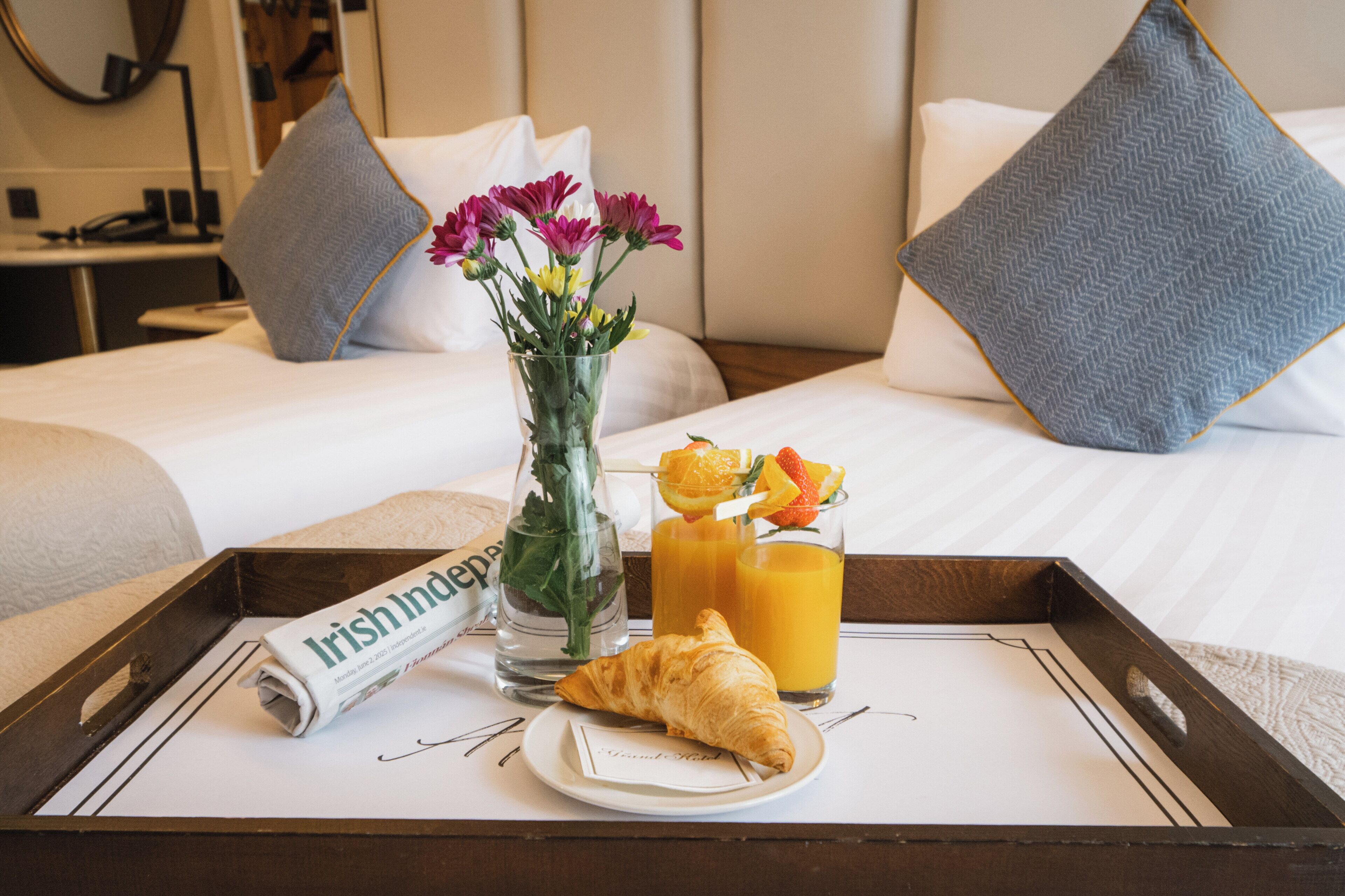 A hotel room with a neatly made bed, a tray with breakfast items including croissant, orange juice, and flowers, and a newspaper.