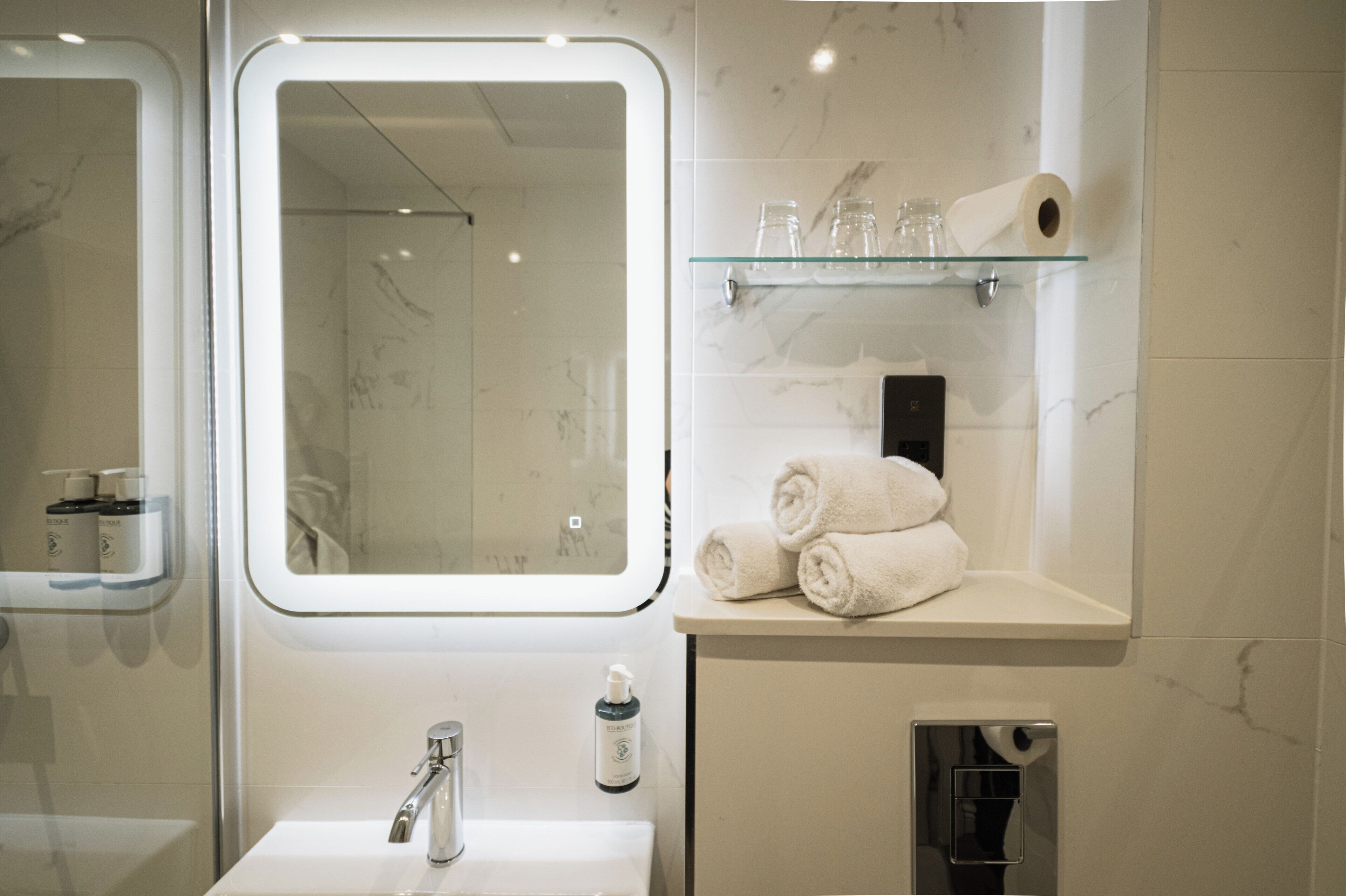 Un baño con un espejo grande, un estante de vidrio con toallas, un dispensador de papel higiénico y un dispensador de jabón para manos.