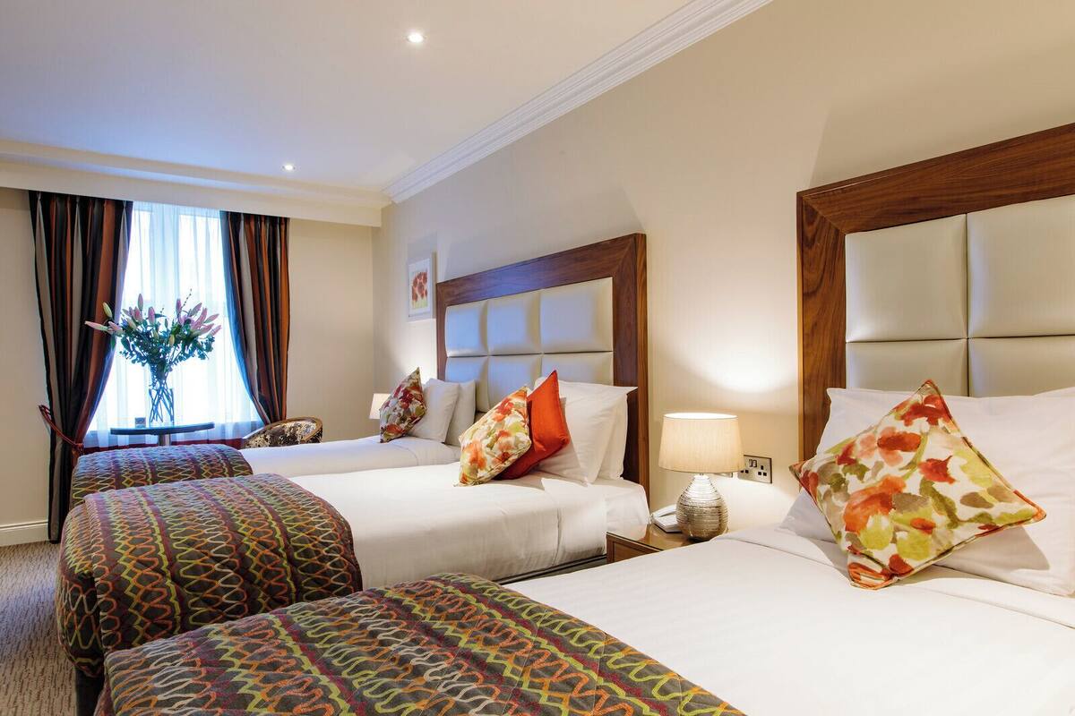 A hotel room with two beds, a nightstand with a lamp, a window with curtains, and a vase with flowers.