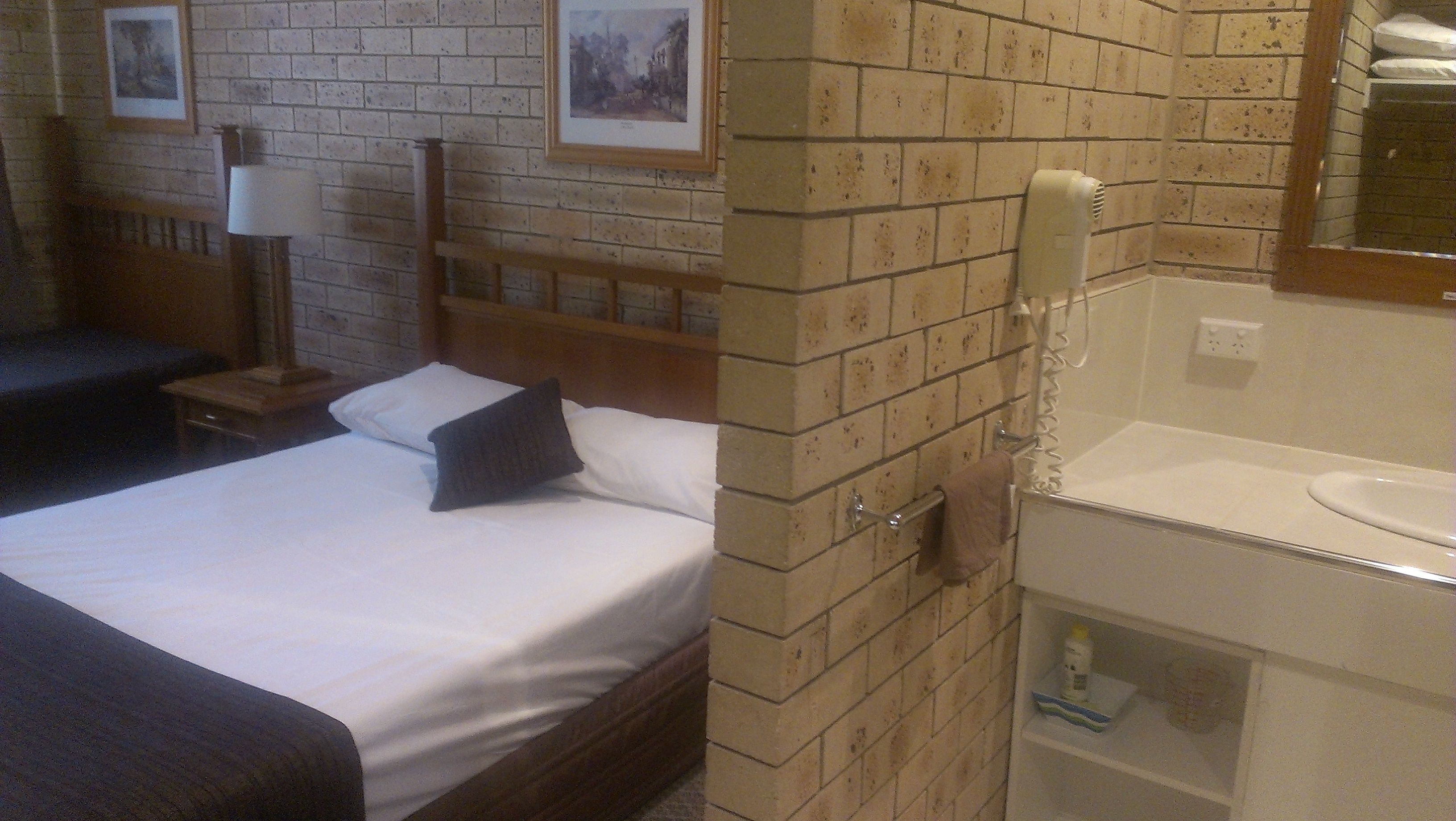 A hotel room with a bed, a nightstand, a lamp, a brick wall, and a bathroom with a sink and mirror.