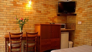 Una habitación pequeña con pared de ladrillo, un escritorio de madera con dos sillas, un televisor montado sobre el escritorio, un microondas y un armario.