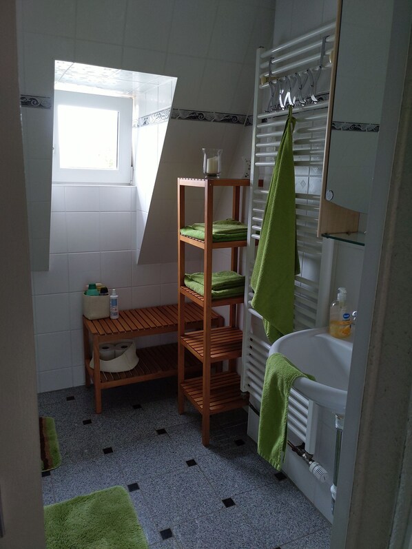 Un baño con un estante de madera con toallas verdes, una mesita con artículos de aseo, una ventana y un lavabo blanco con una toalla verde colgada en el radiador.
