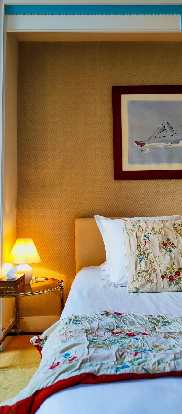 A hotel room with a bed, a bedside table with a lamp, a framed artwork on the wall, and a floral-patterned pillow.