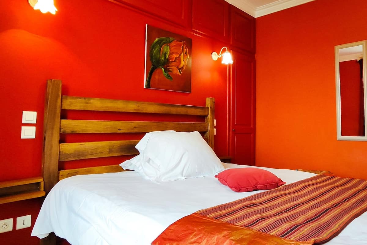 Una habitación con cabecera de madera, ropa de cama blanca, manta a rayas rojas y anaranjadas, y una pared roja con un cuadro.