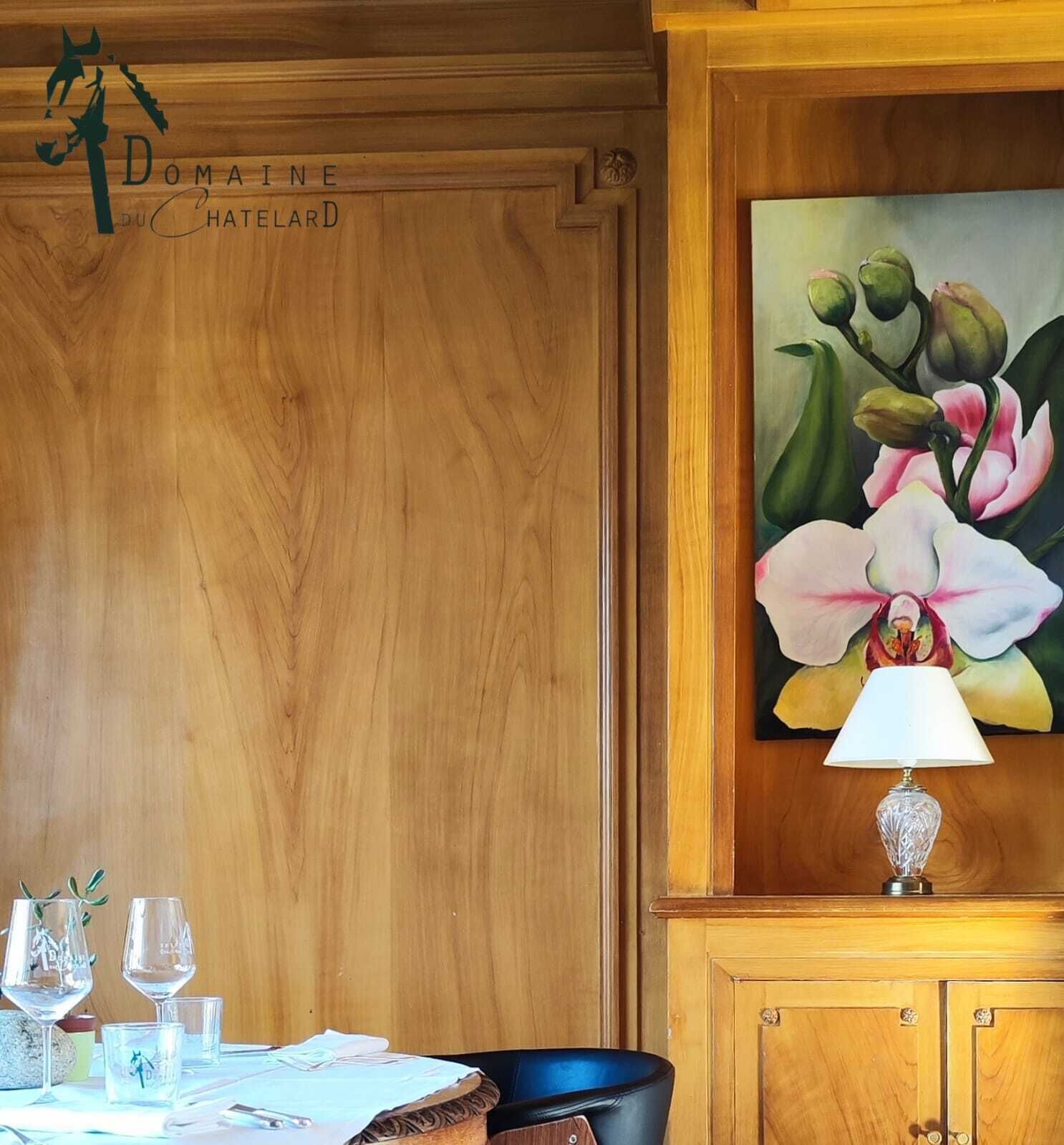 A wooden-paneled room with a painting of flowers, a table set with glasses and a lamp, and a sign reading "Domaine du Chatelier".