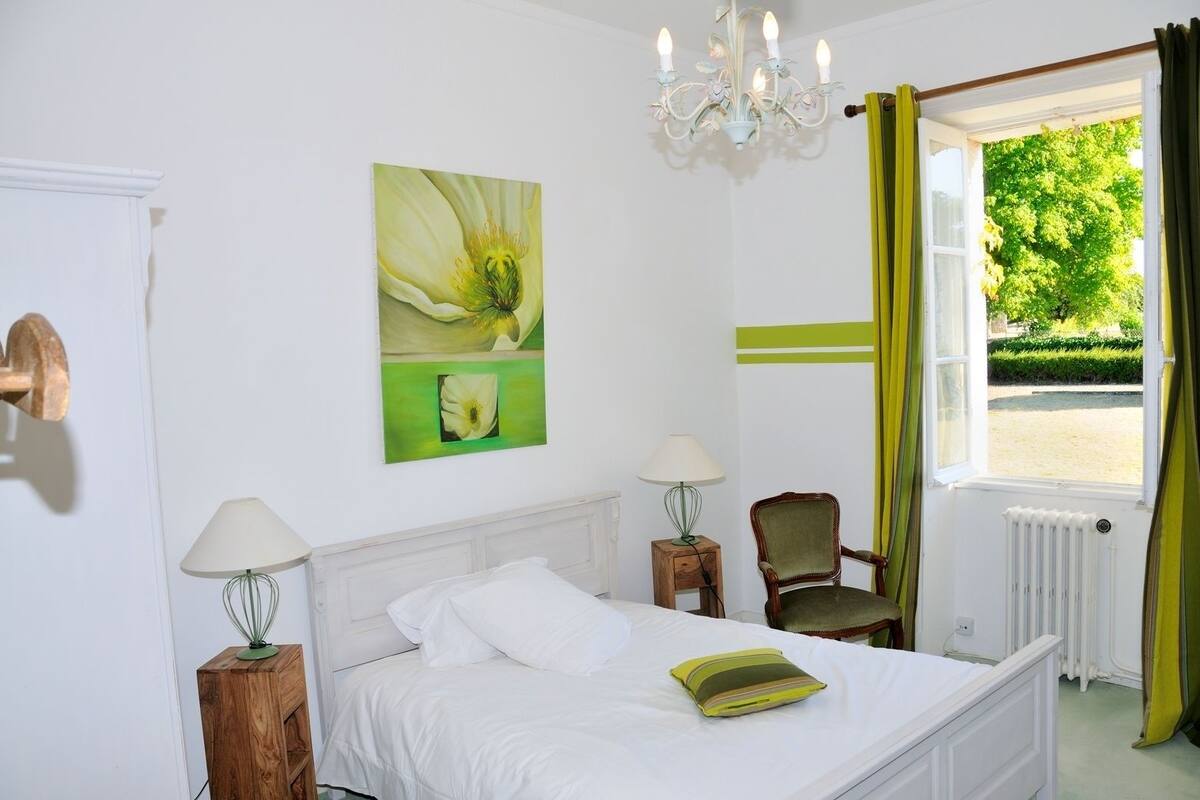 Un dormitorio con cama, dos mesitas de noche, una silla, una araña, ventana con cortinas y un cuadro en la pared.