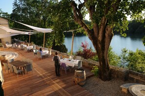 An outdoor dining area with tables and chairs, overlooking a lake, surrounded by trees and a stone wall.