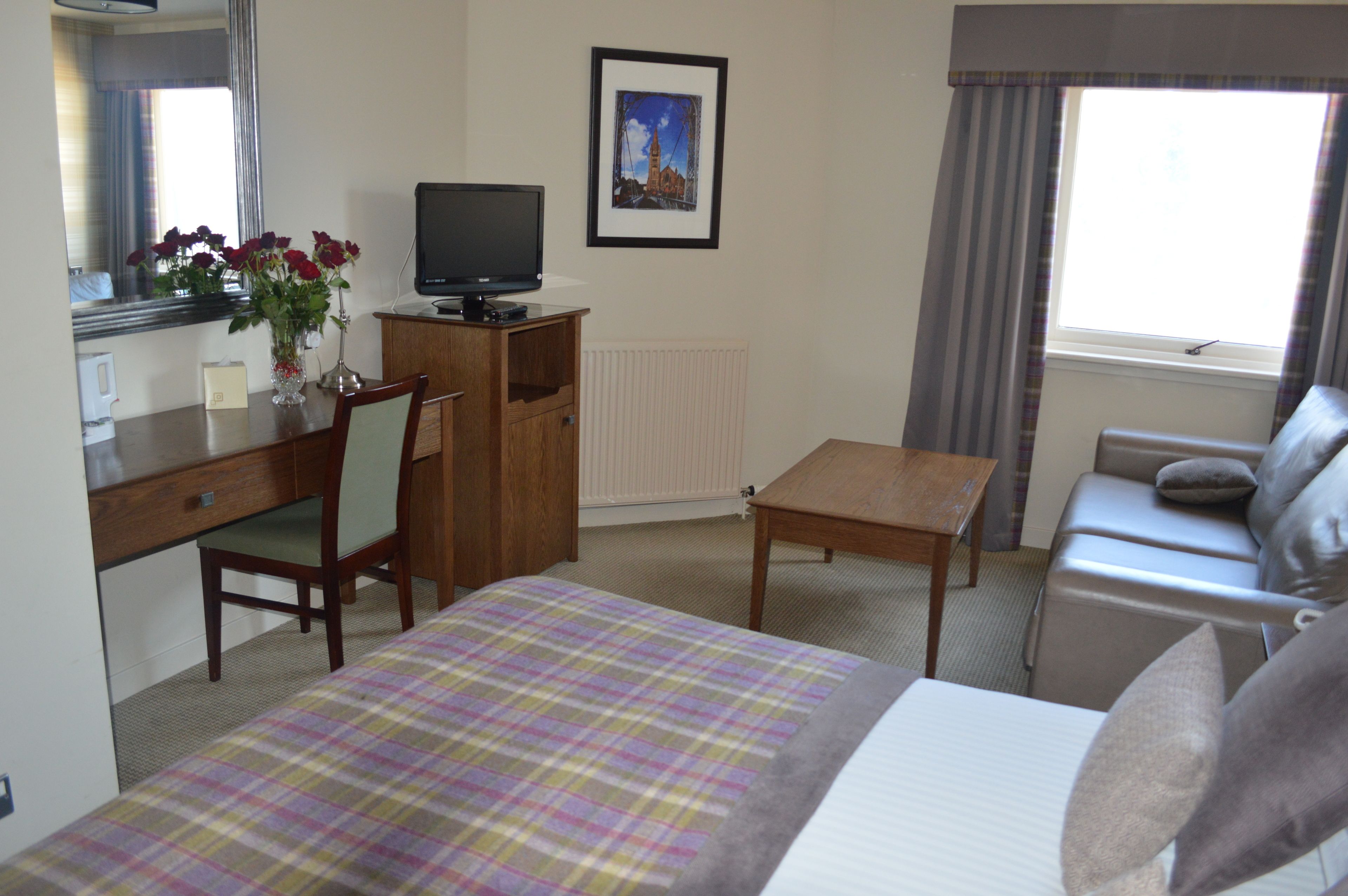 A hotel room with a bed, a desk with a computer, a chair, a sofa, a small table, a mirror, a picture on the wall, and a window with curtains.
