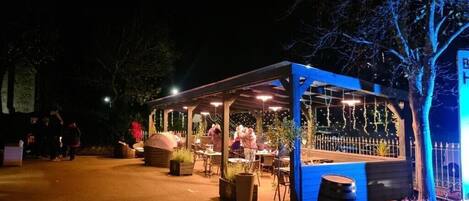 An outdoor dining area with tables and chairs, surrounded by trees and a fence.
