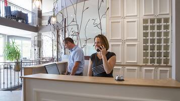 A reception area with a wooden counter, a man standing behind it, and a woman talking on the phone. There are white cabinets with numbered drawers and a balcony with a railing in the background.
