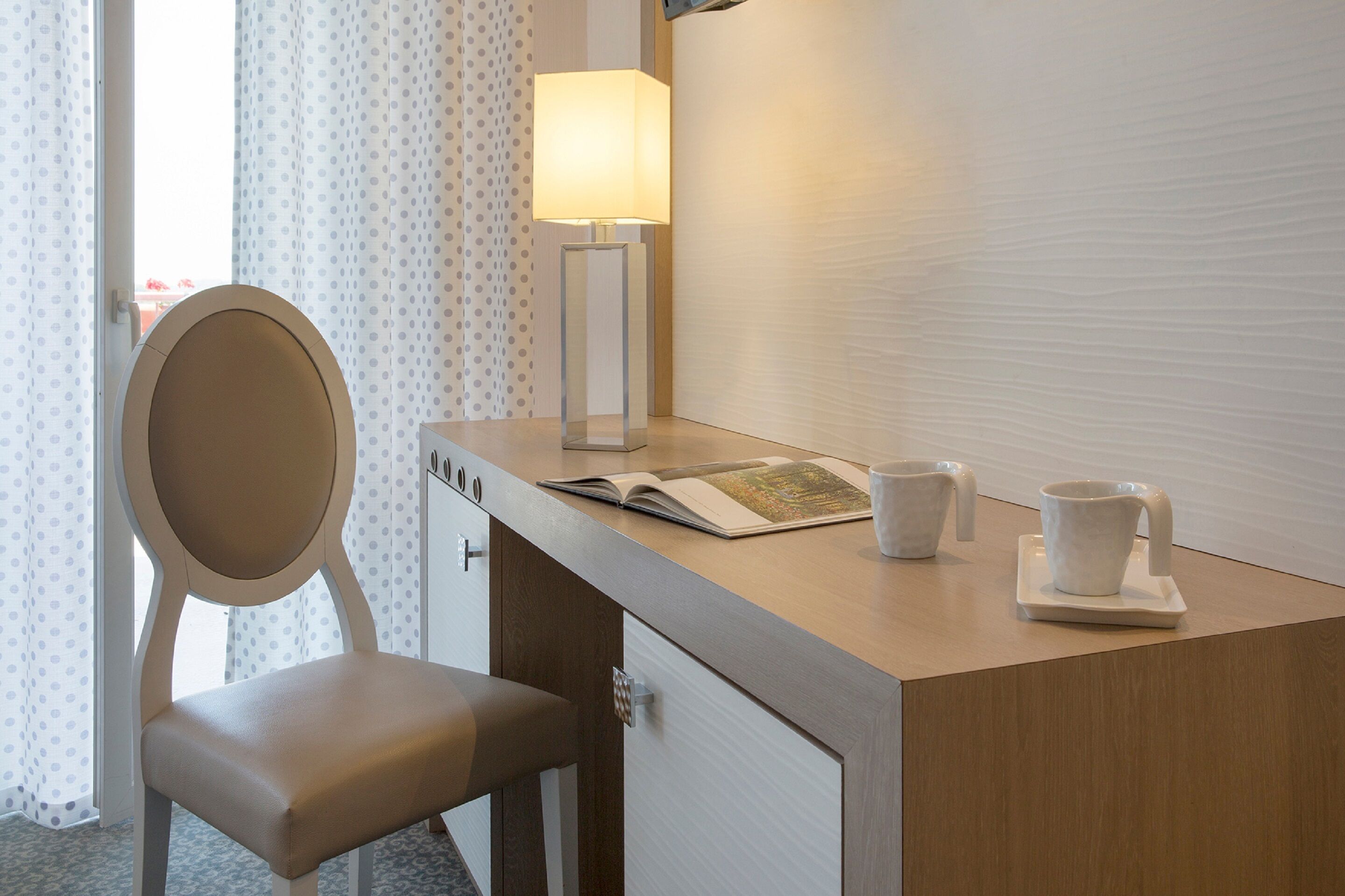 Una habitación de hotel con un escritorio de madera, una silla, una lámpara, un libro y dos tazas sobre una bandeja.