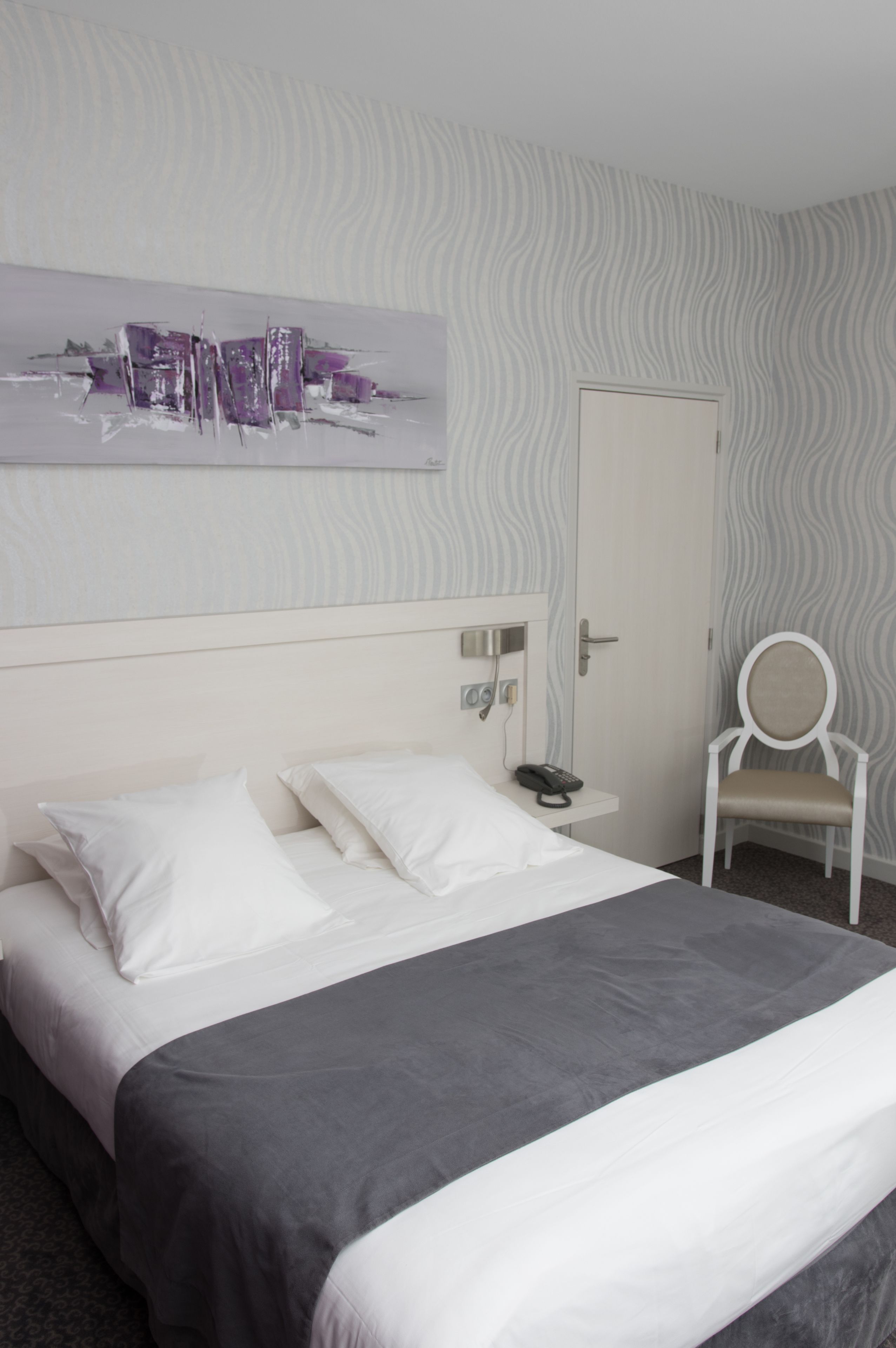 A hotel room with a bed, a bedside table with a phone, a chair, and a door.