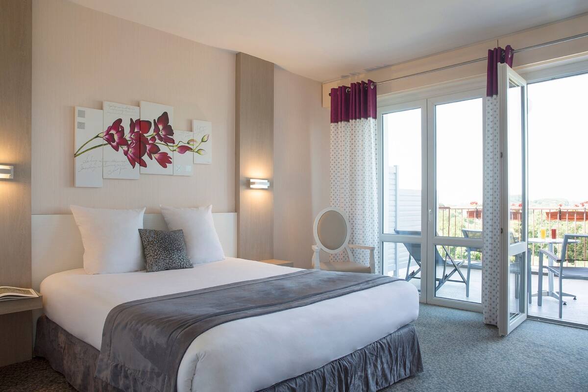 A hotel room with a large bed, a bedside table, a chair, a balcony with a view, and artwork on the wall.