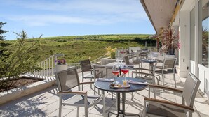 A terrace with outdoor seating, a view of a vineyard, and a building with a sloped roof.