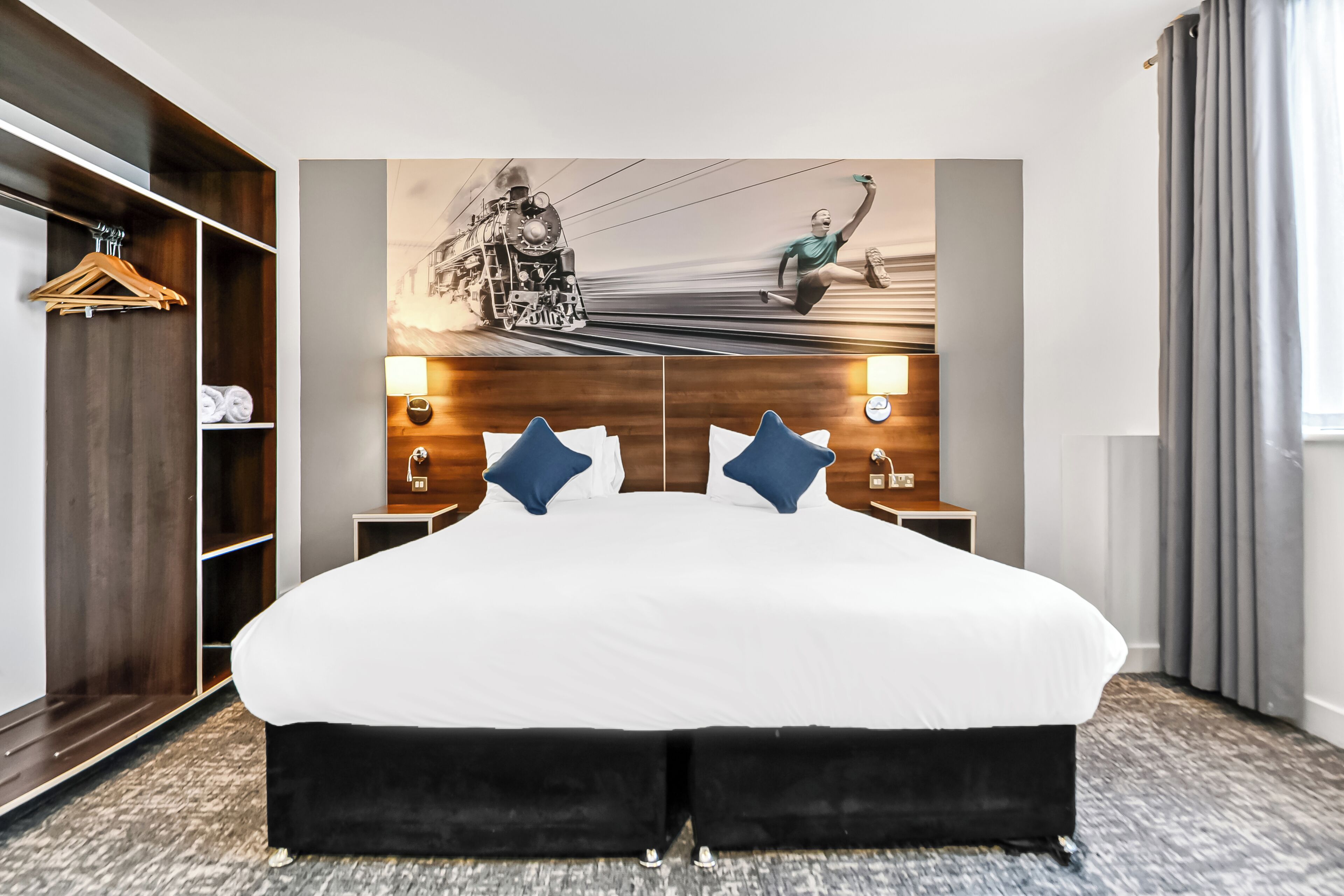 Quarto de hotel com uma cama grande, um guarda-roupa de madeira, uma luminária na parede e um quadro de um trem.