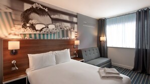 A hotel room with a large bed, a grey armchair, a small table with a lamp, and a wall decoration featuring glasses and popcorn. - Heeton Concept Hotel City Centre Liverpool (Liverpool)