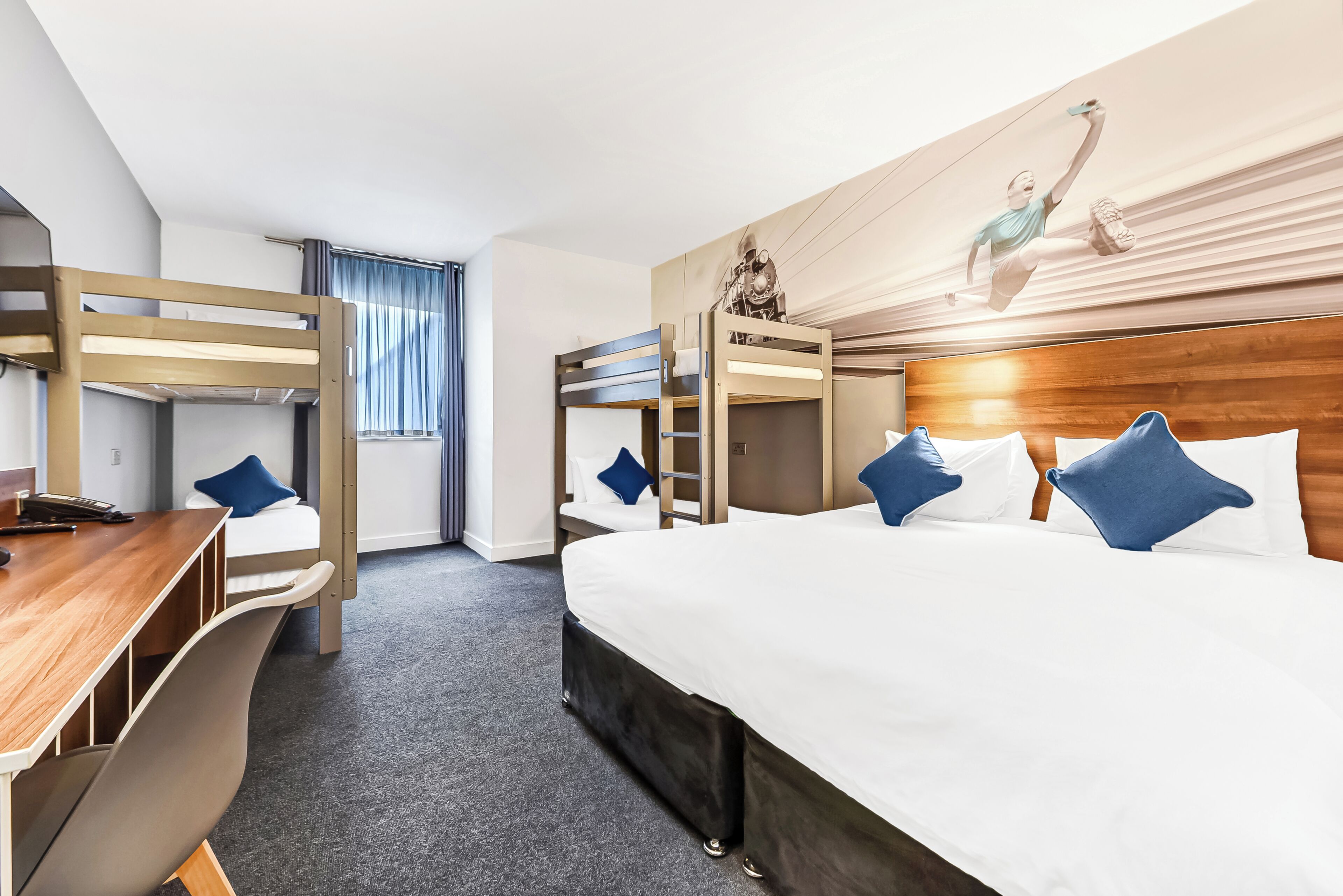 A hotel room with bunk beds, a desk, and a bed with pillows.