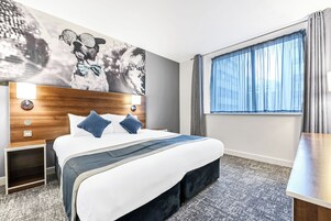 Una habitación de hotel con una cama, una mesita de noche, una lámpara de pared, una ventana con cortinas y un cuadro abstracto de gran tamaño.