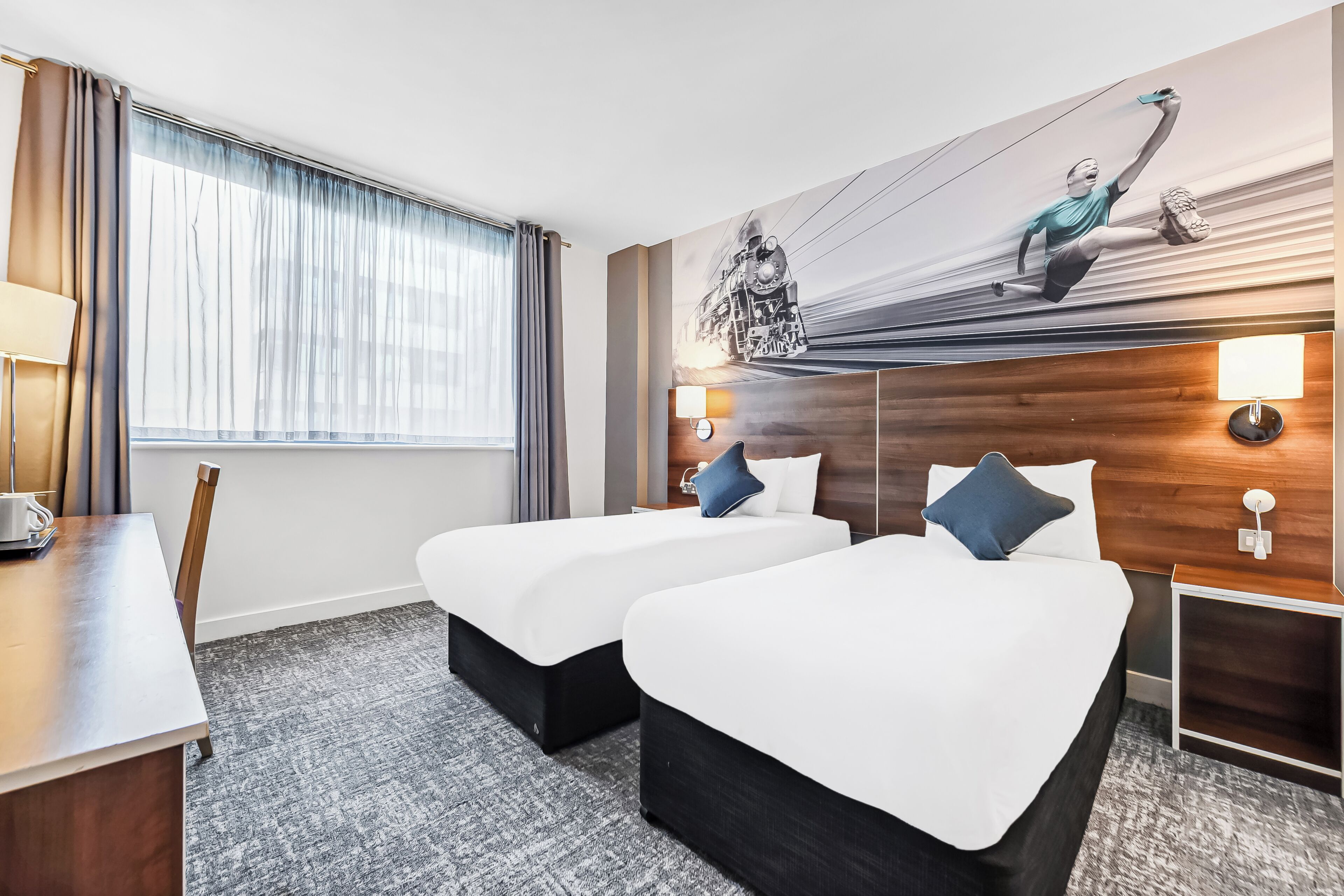 A hotel room with two beds, a desk, a chair, a window with curtains, and a wall mural.