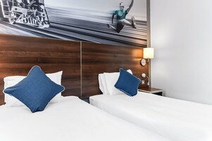 A hotel room with two beds, each with a white mattress and a blue pillow. There is a wooden headboard, a nightstand with a lamp, and a wall-mounted light fixture.