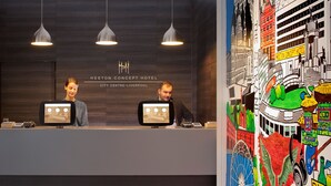 A hotel reception desk with two staff members, a modern interior with pendant lighting, and a colorful mural on the wall. - Heeton Concept Hotel City Centre Liverpool (Liverpool)