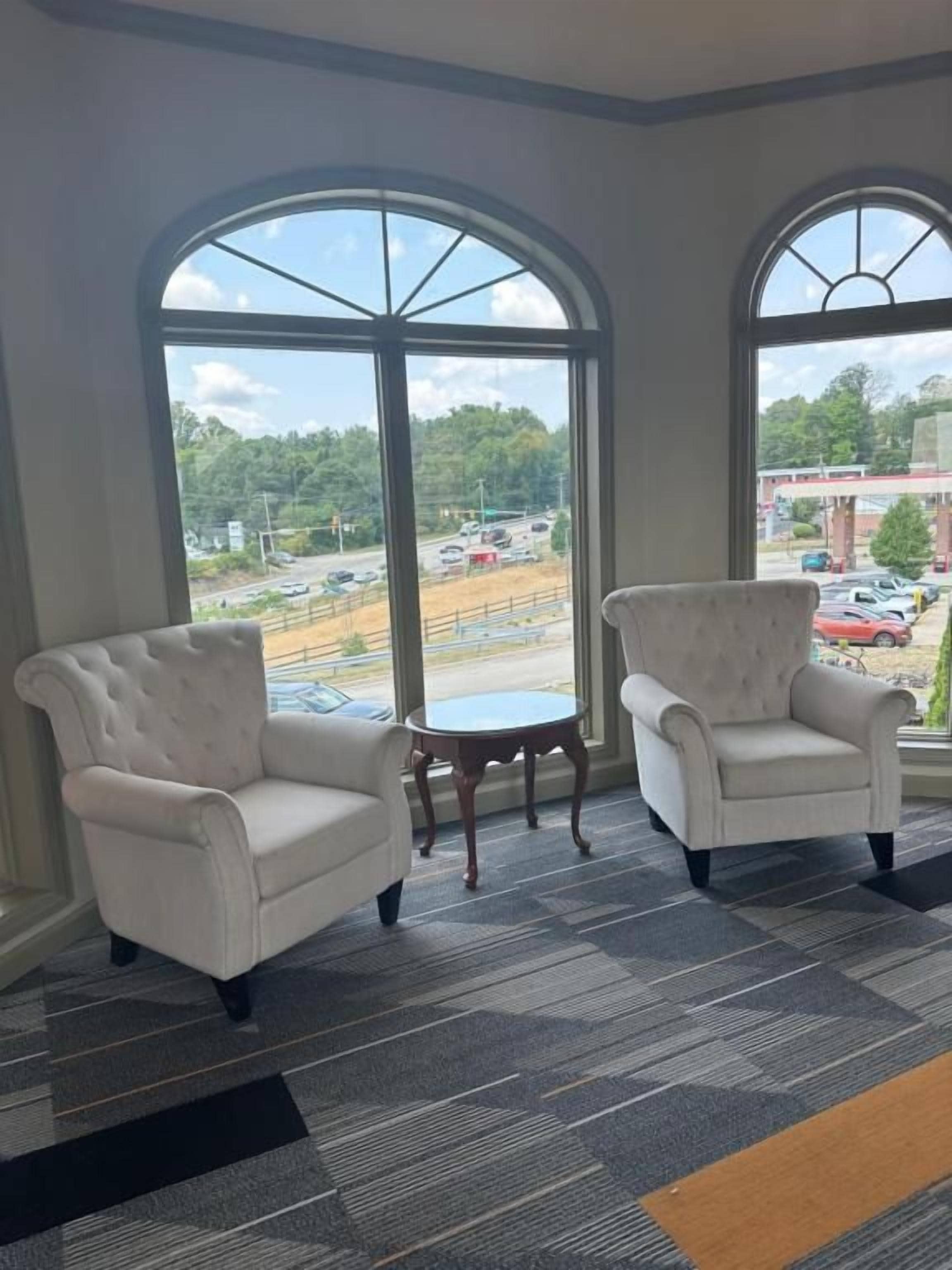 A room with two white armchairs and a small round table, large arched windows with a view of a parking lot and trees.