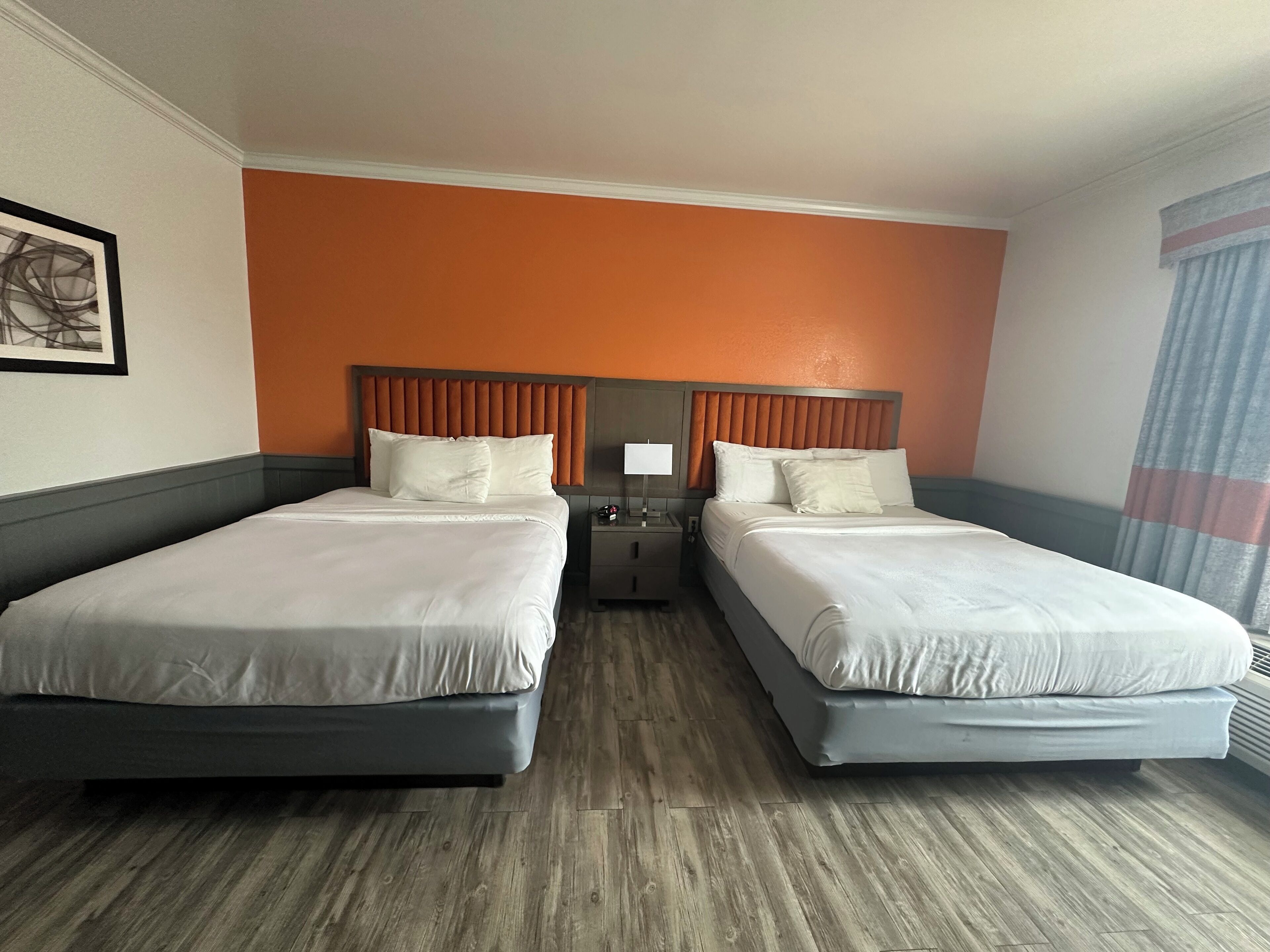 A hotel room with two beds, a wooden floor, an orange accent wall, and a framed picture on the wall.