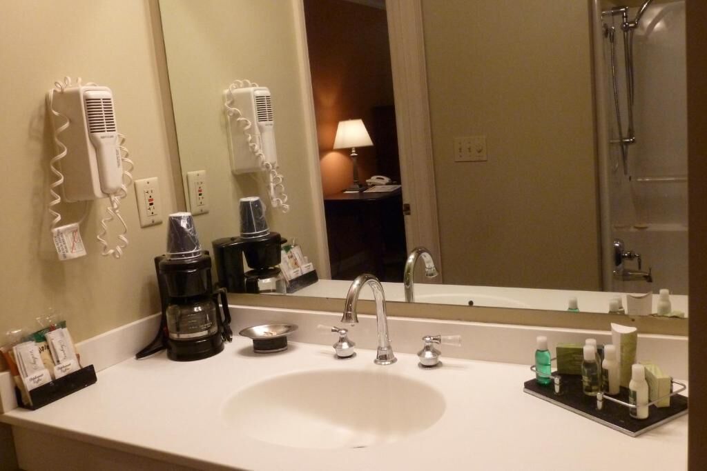 A hotel bathroom with a sink, two hair dryers, a coffee maker, and toiletries on the counter.