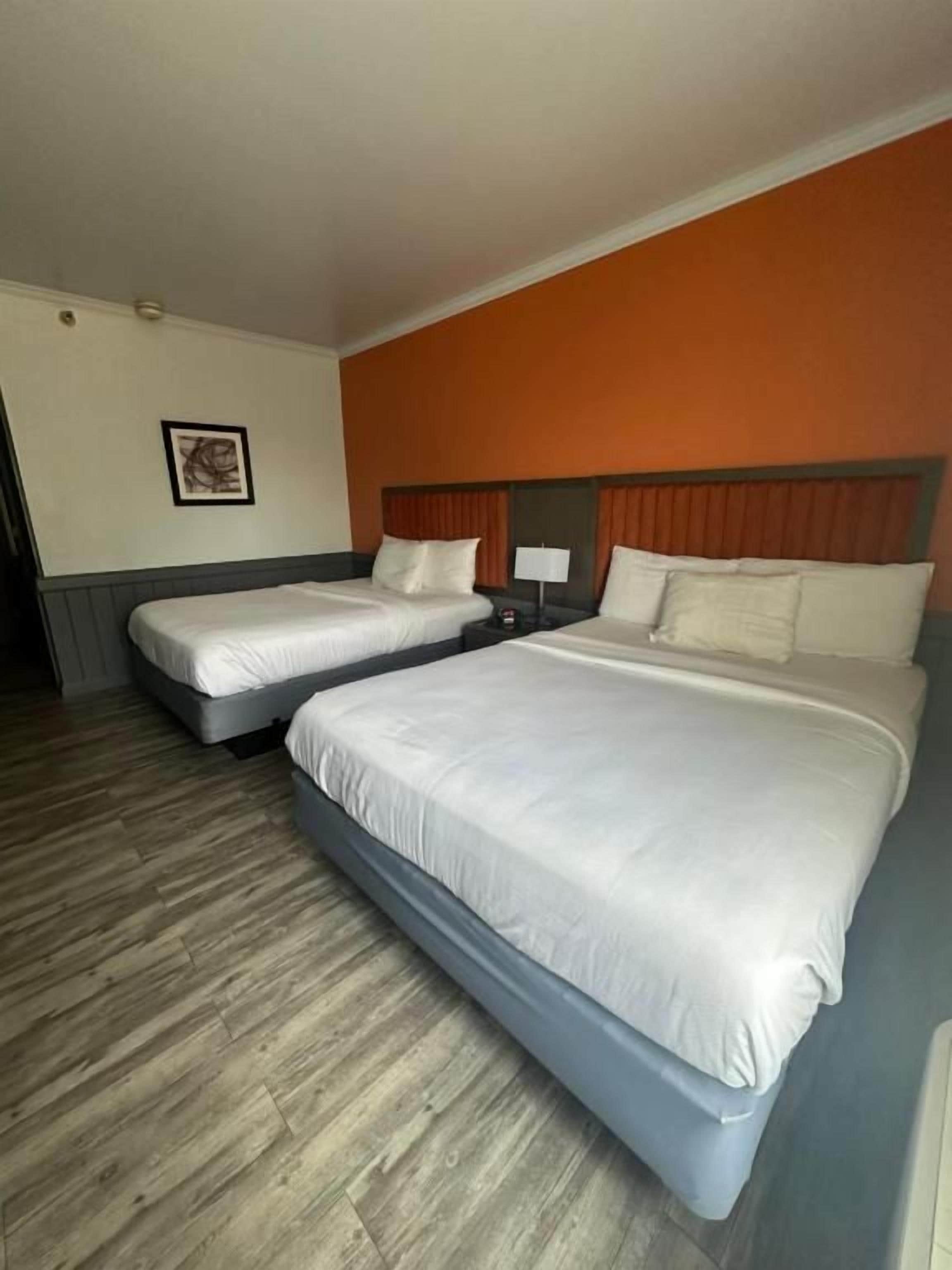 A hotel room with two beds, a wooden floor, and a single wall with a framed picture.