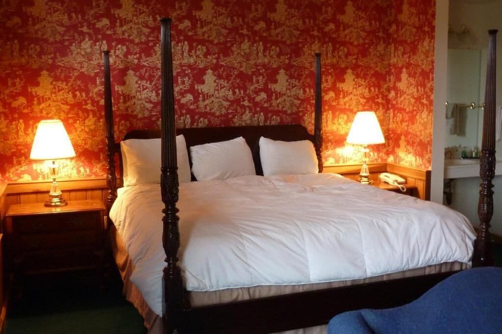 A four-poster bed with white linens, two bedside tables with lamps, and a floral wallpaper pattern.
