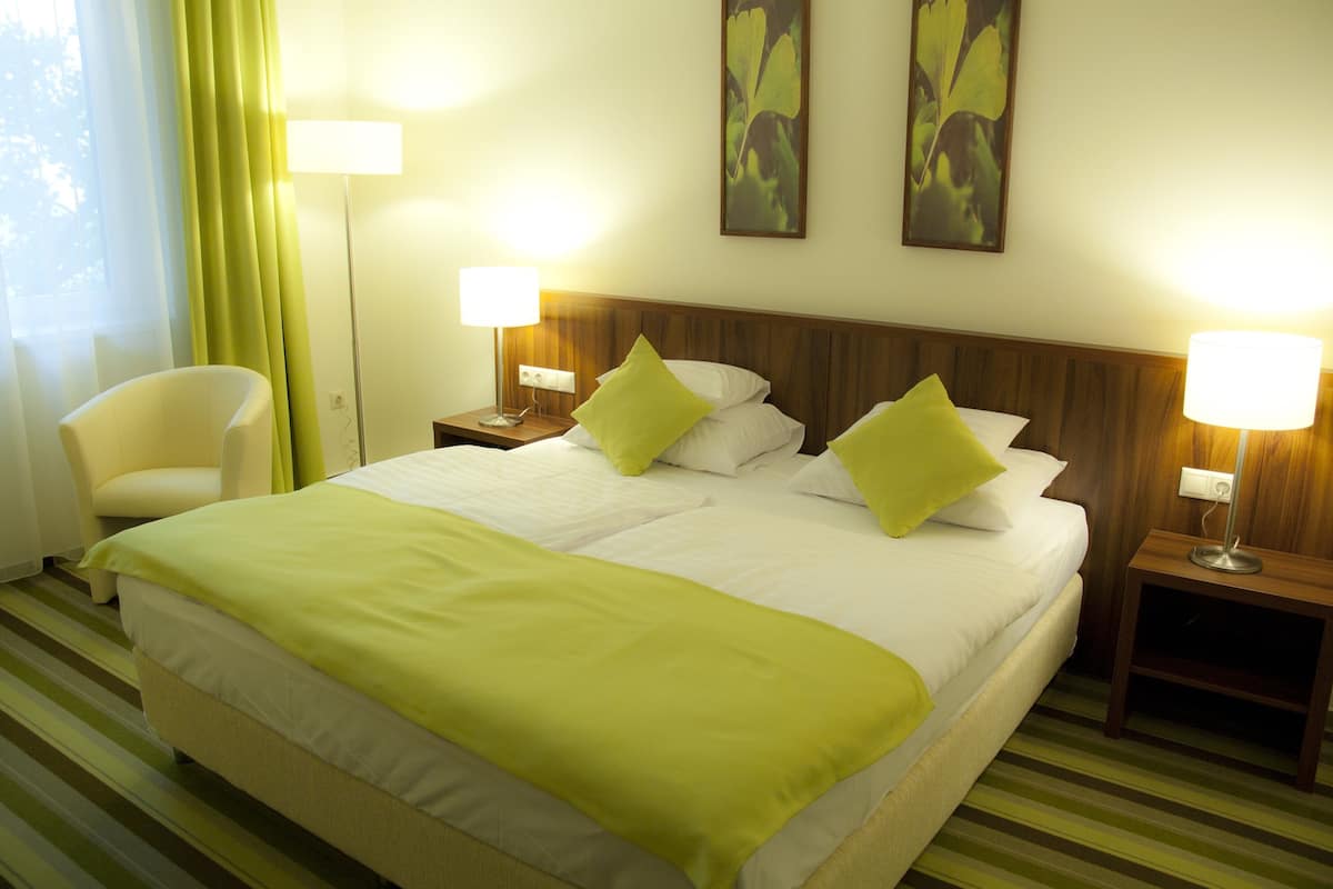 Quarto de hotel com uma cama grande, duas luminárias de cabeceira, uma cadeira e uma janela com cortinas.