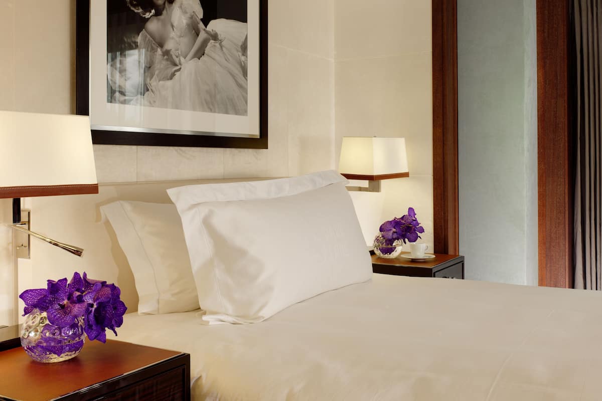 A hotel room with a bed, two bedside tables, a framed picture on the wall, and a vase with purple flowers on the nightstand.