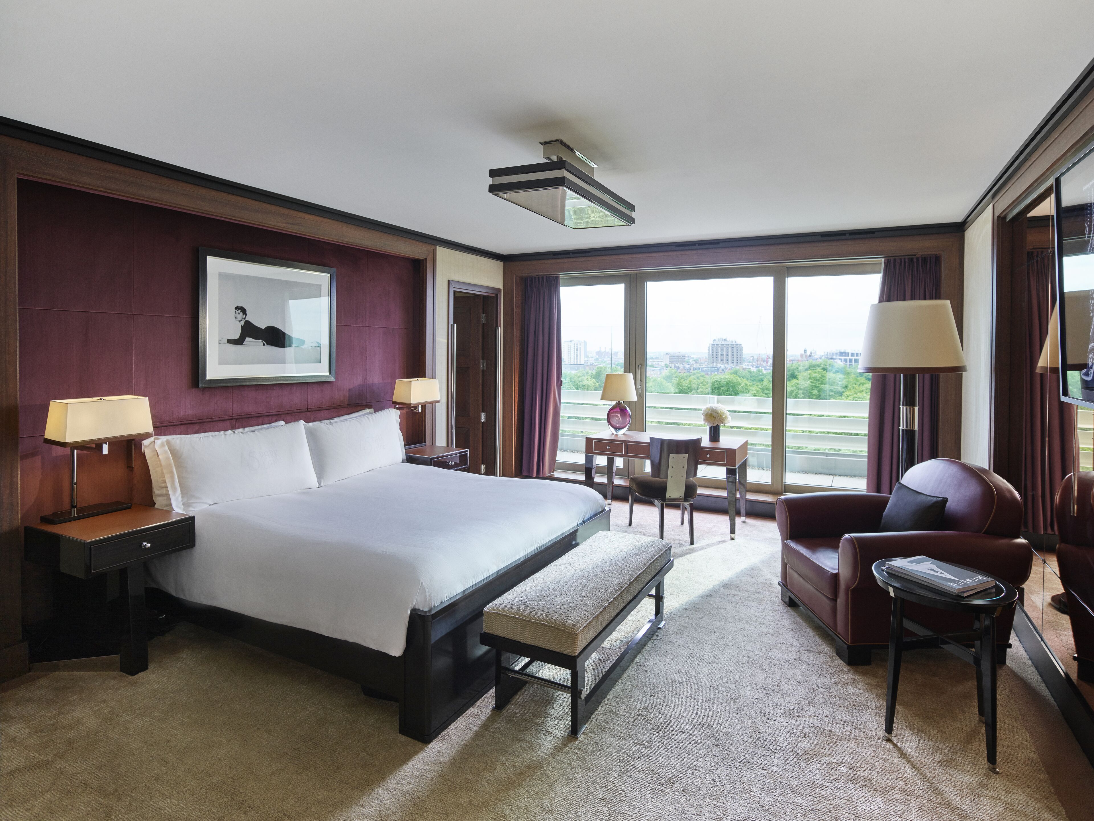 Habitación de hotel con una cama grande, un escritorio, una silla y vistas de la ciudad a través de amplias ventanas.