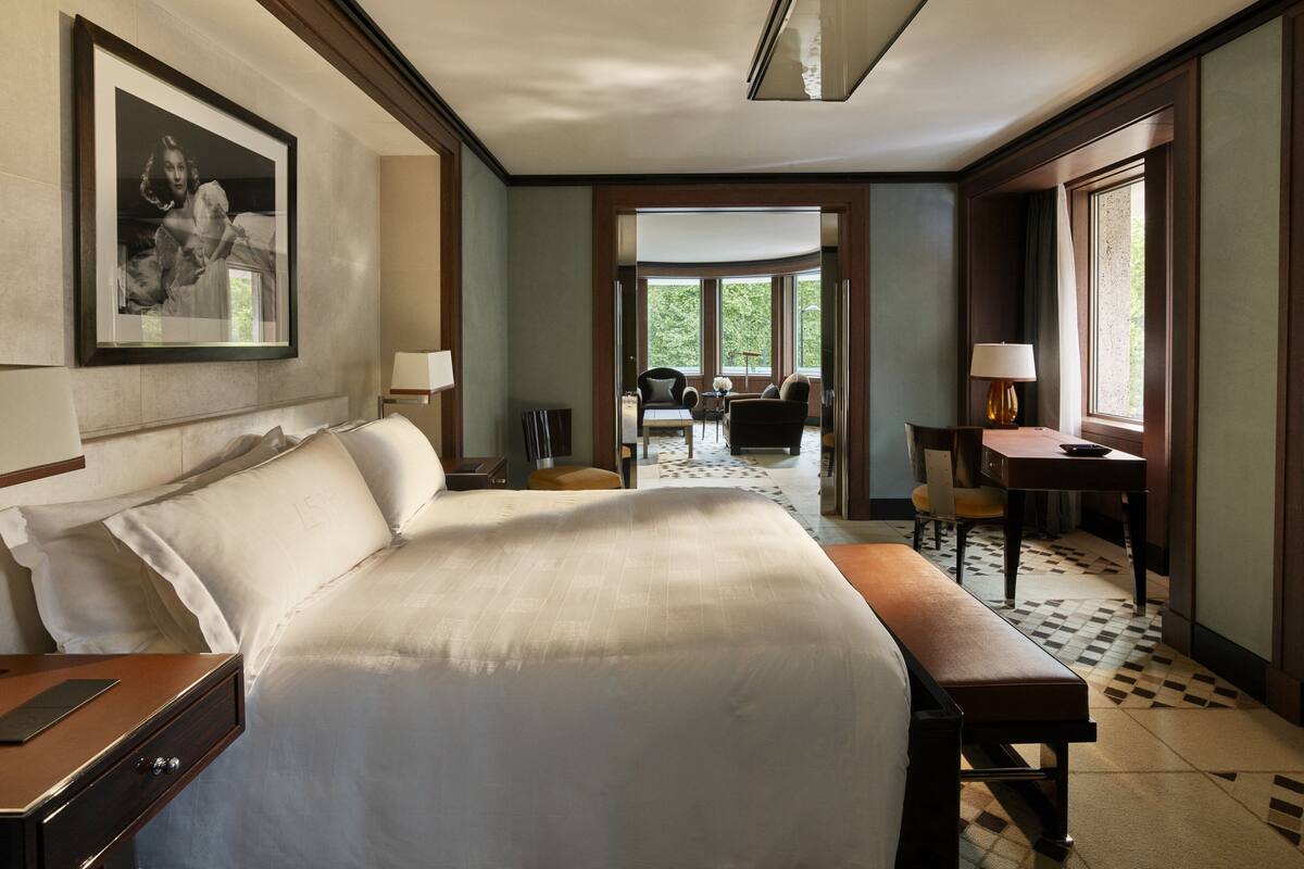 A hotel room with a large bed, a desk, a chair, and a dining area with a view of the outdoors.