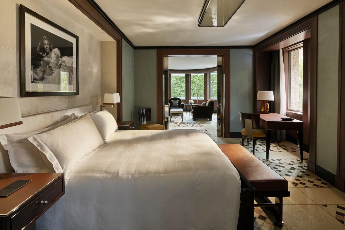 A hotel room with a large bed, a desk, a chair, and a dining area visible through an open door.