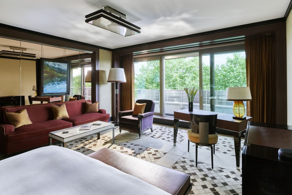 A hotel room with a large bed, a red sofa, a coffee table, a TV, and a window with a view of greenery.