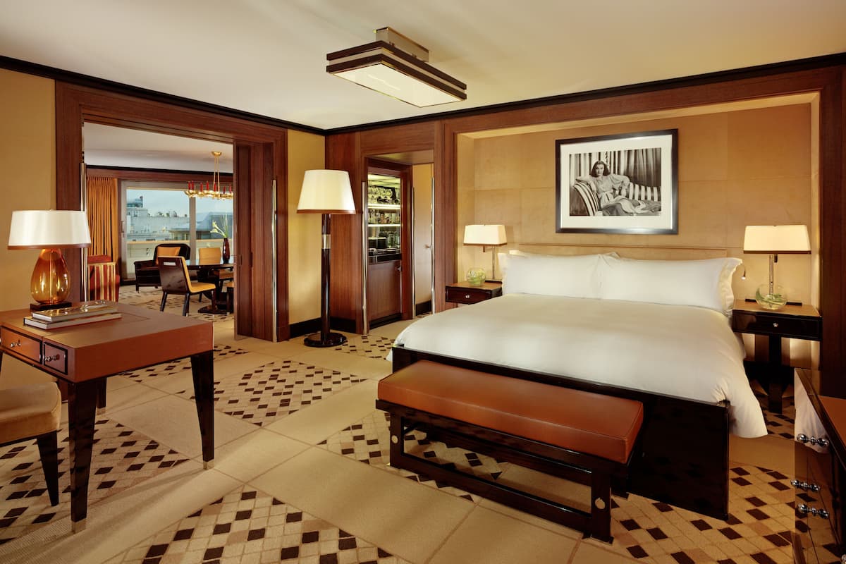 A hotel room with a large bed, a desk, a chair, a bench, and a dining area with a table and chairs.