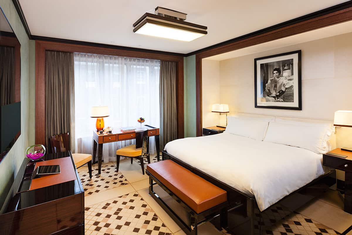A hotel room with a large bed, a desk with a chair, a bench, a desk lamp, and a framed picture on the wall.