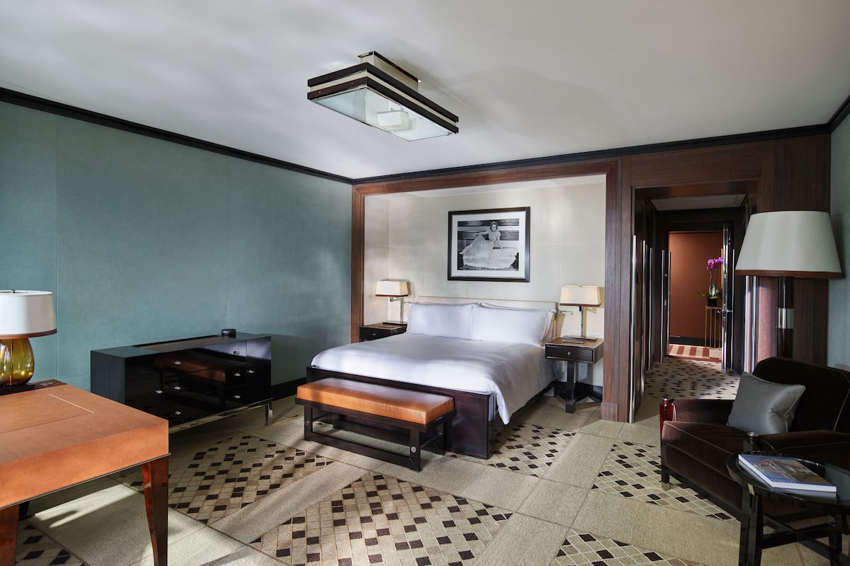 A hotel room with a large bed, a desk, a chair, and a sofa. There is a picture on the wall and a lamp on the desk.
