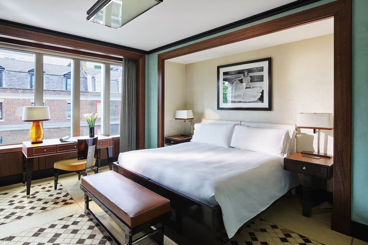 A hotel room with a large bed, a bench, a desk, and a window with a view of buildings.