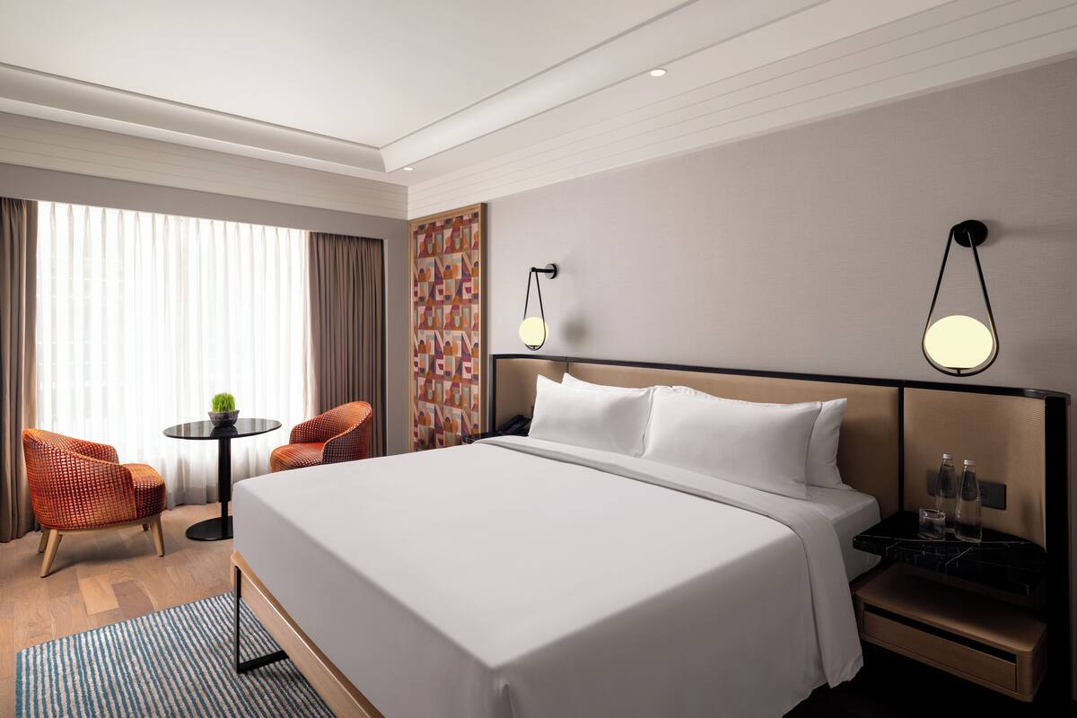 Habitación de hotel con una cama grande, una mesita con una silla, una mesita de noche con lámpara y un panel decorativo en la pared.