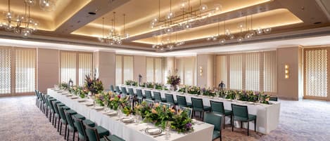 Un salón de conferencias con una larga mesa dispuesta para un evento formal, rodeado de sillas, una alfombra estampada y luminarias modernas.